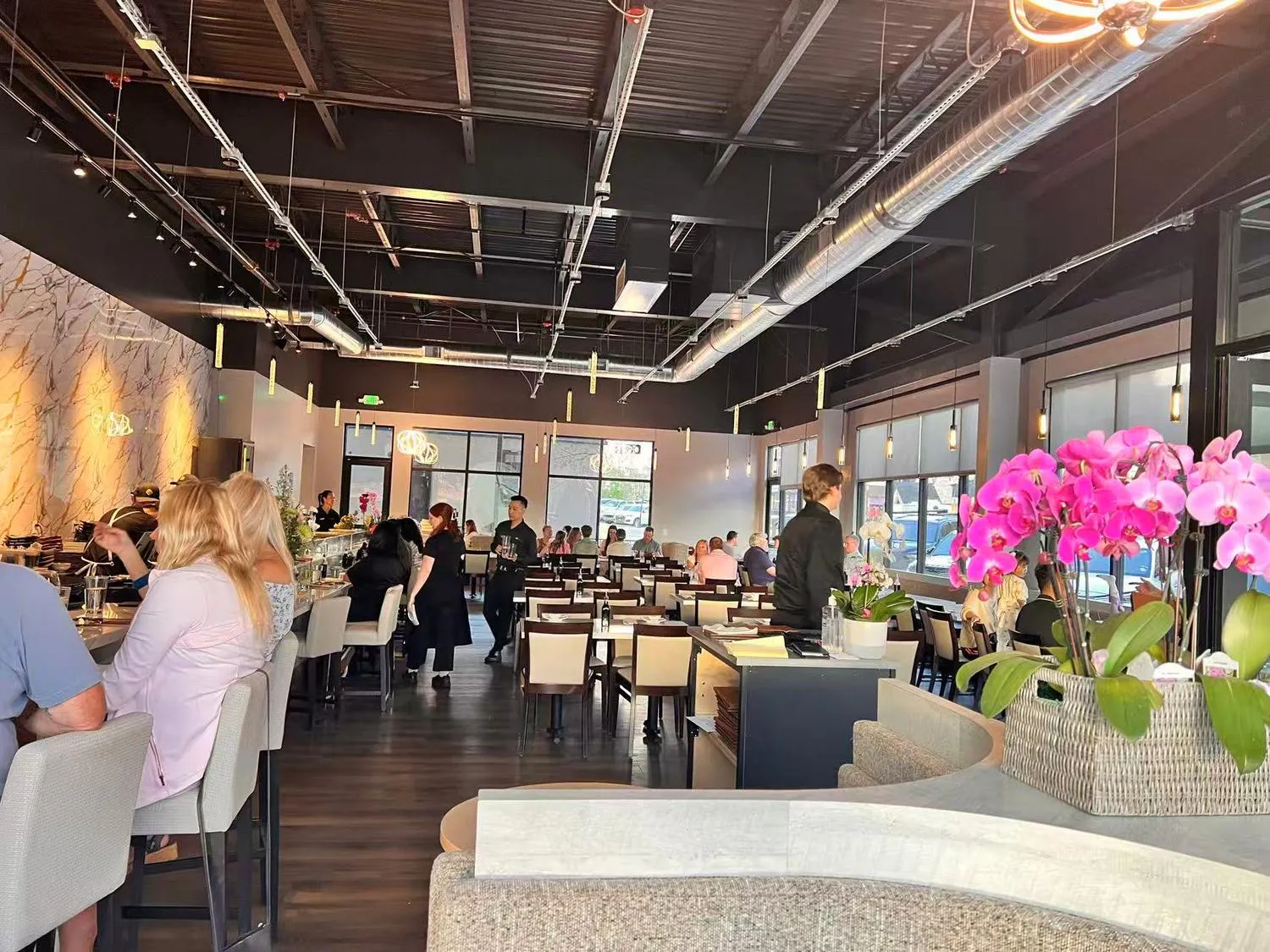 Modern restaurant dining room with guests, bar seating, large windows, and pink orchids